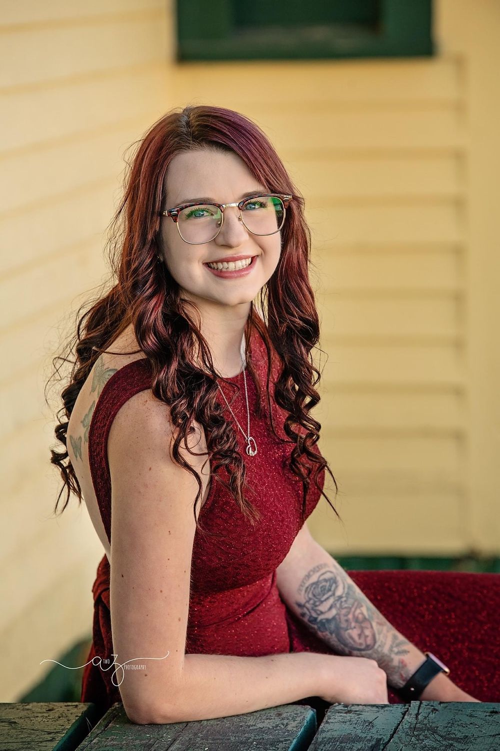 A portrait of someone wearing glasses and a burgundy dress against a yellow wall background.