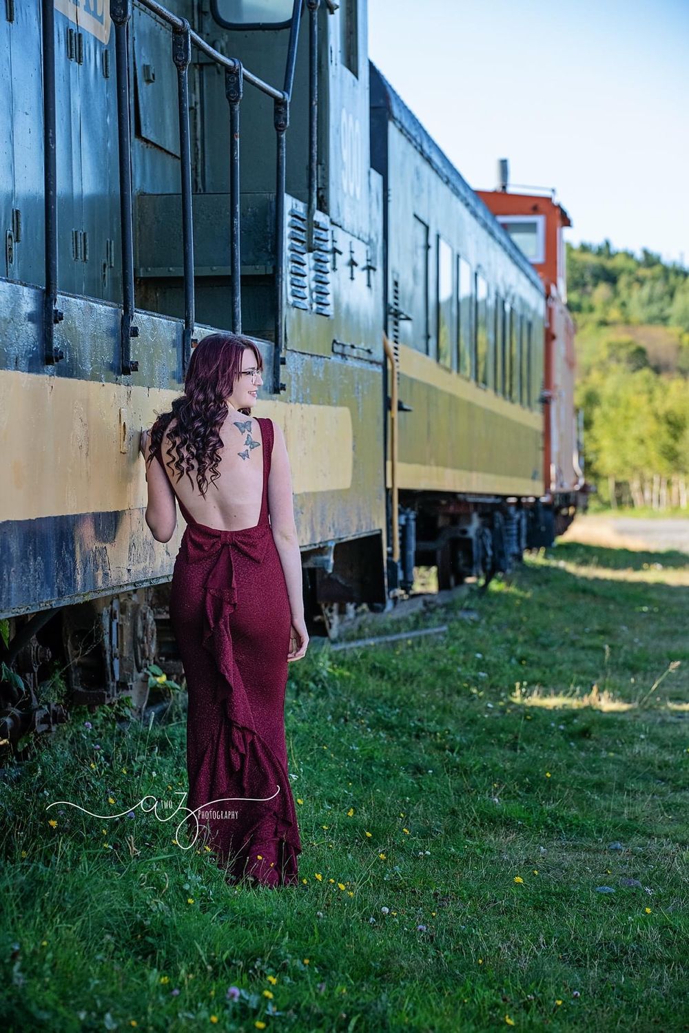 A person in a burgundy dress poses beside a vintage train car in natural lighting.