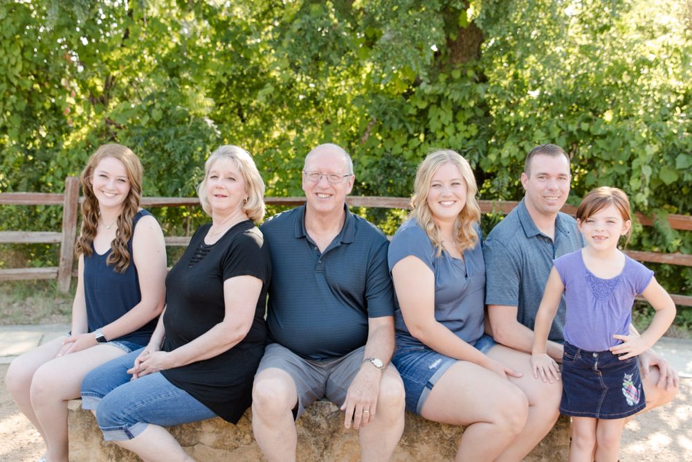 Casual family portrait seated together on wooden bench outdoors with greenery in background.