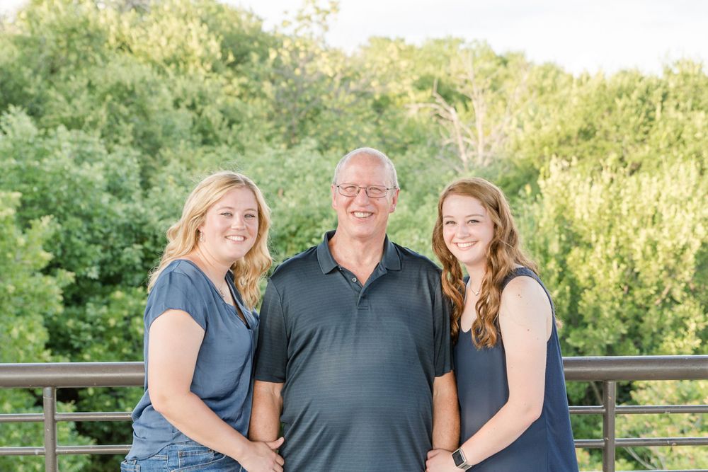 Three people pose together on outdoor deck with sunlit foliage in background.