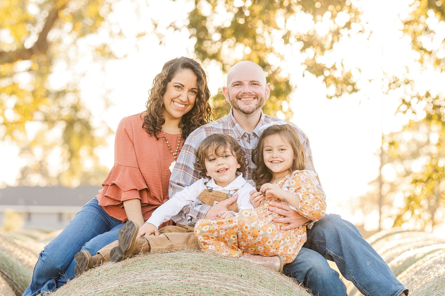 Family of four sitting together on hay bales during golden hour sunset photo session.