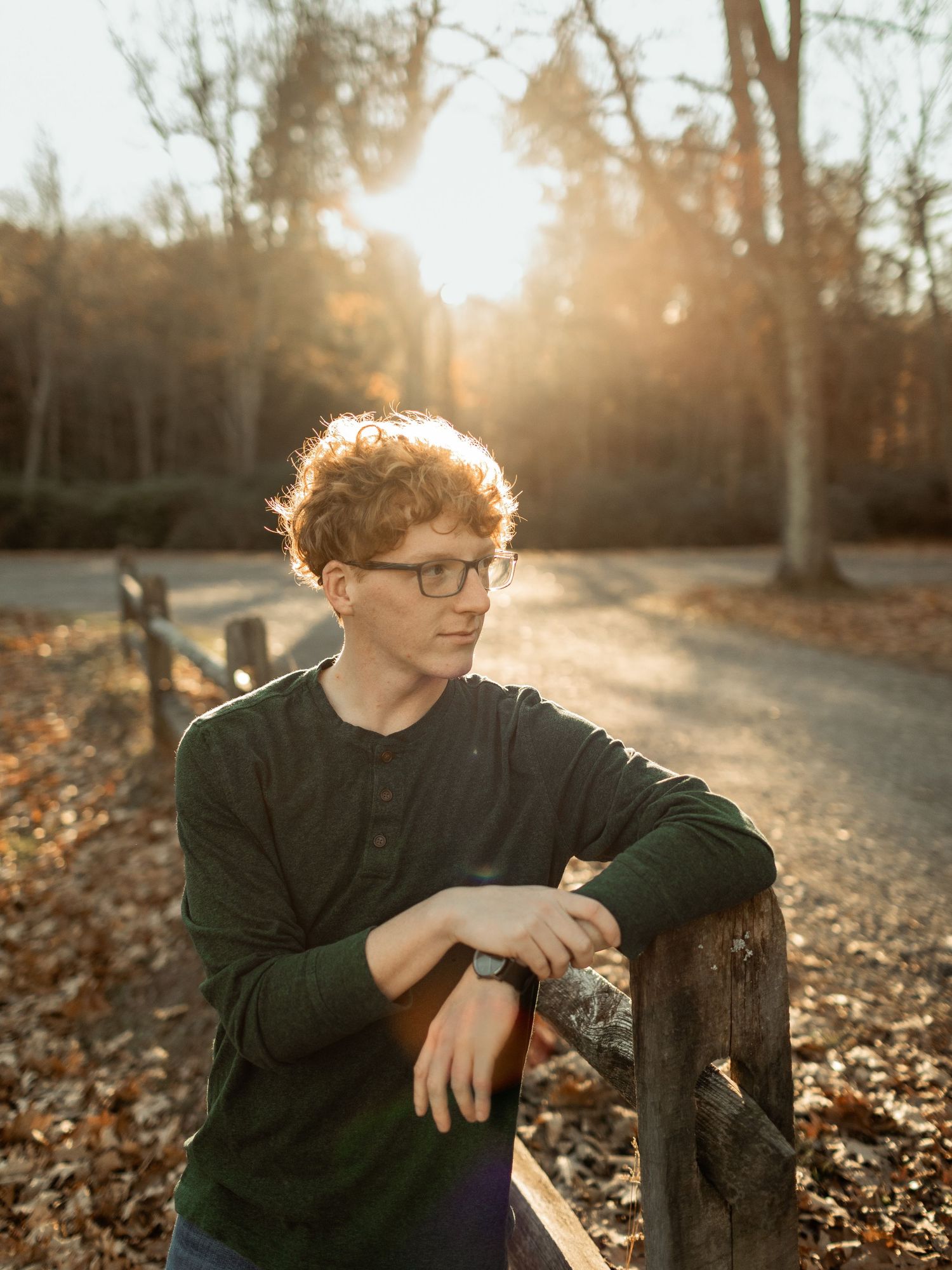 Backlit portrait of young person in glasses leaning on wooden fence during sunset.
