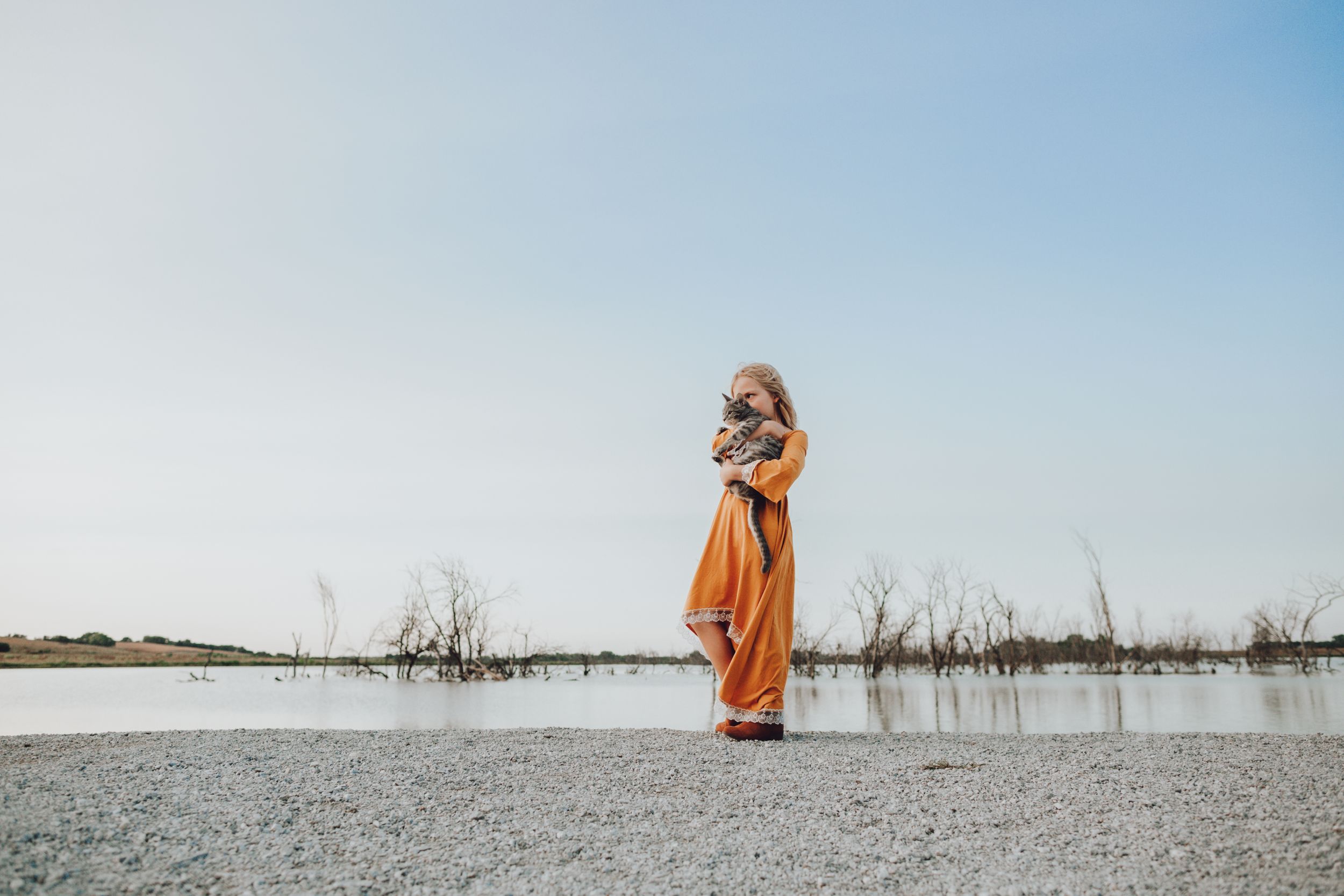 An orange-clad silhouette stands near a tranquil lake with dead trees.
