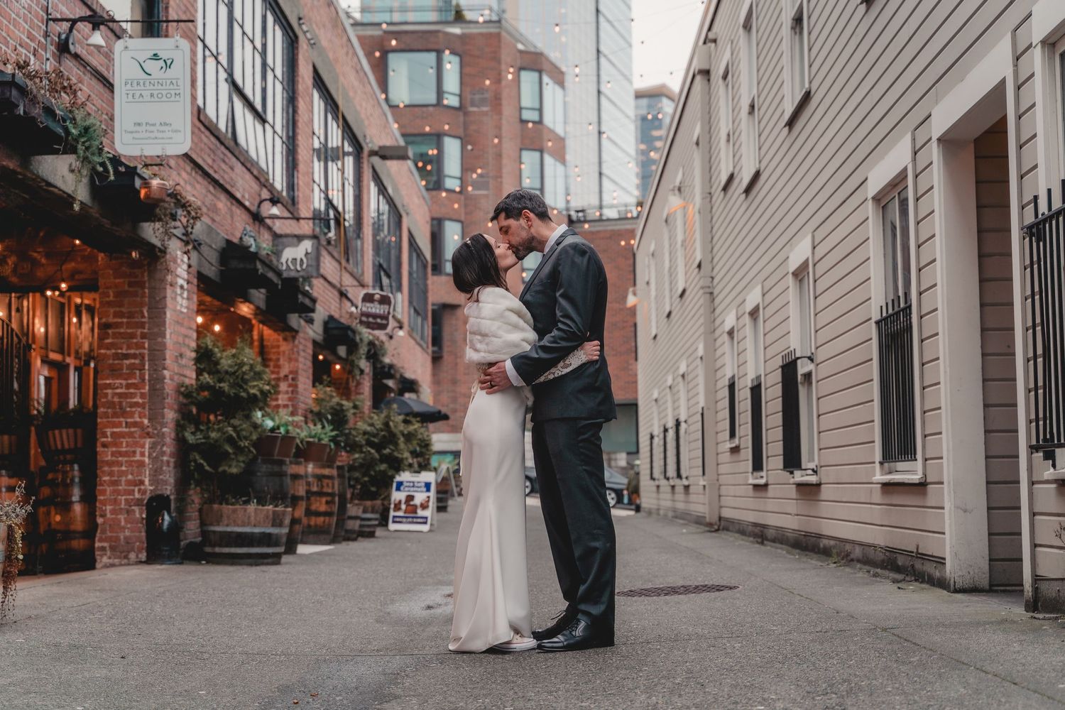 A couple shares an intimate moment in an alley between historic brick buildings decorated with greenery.