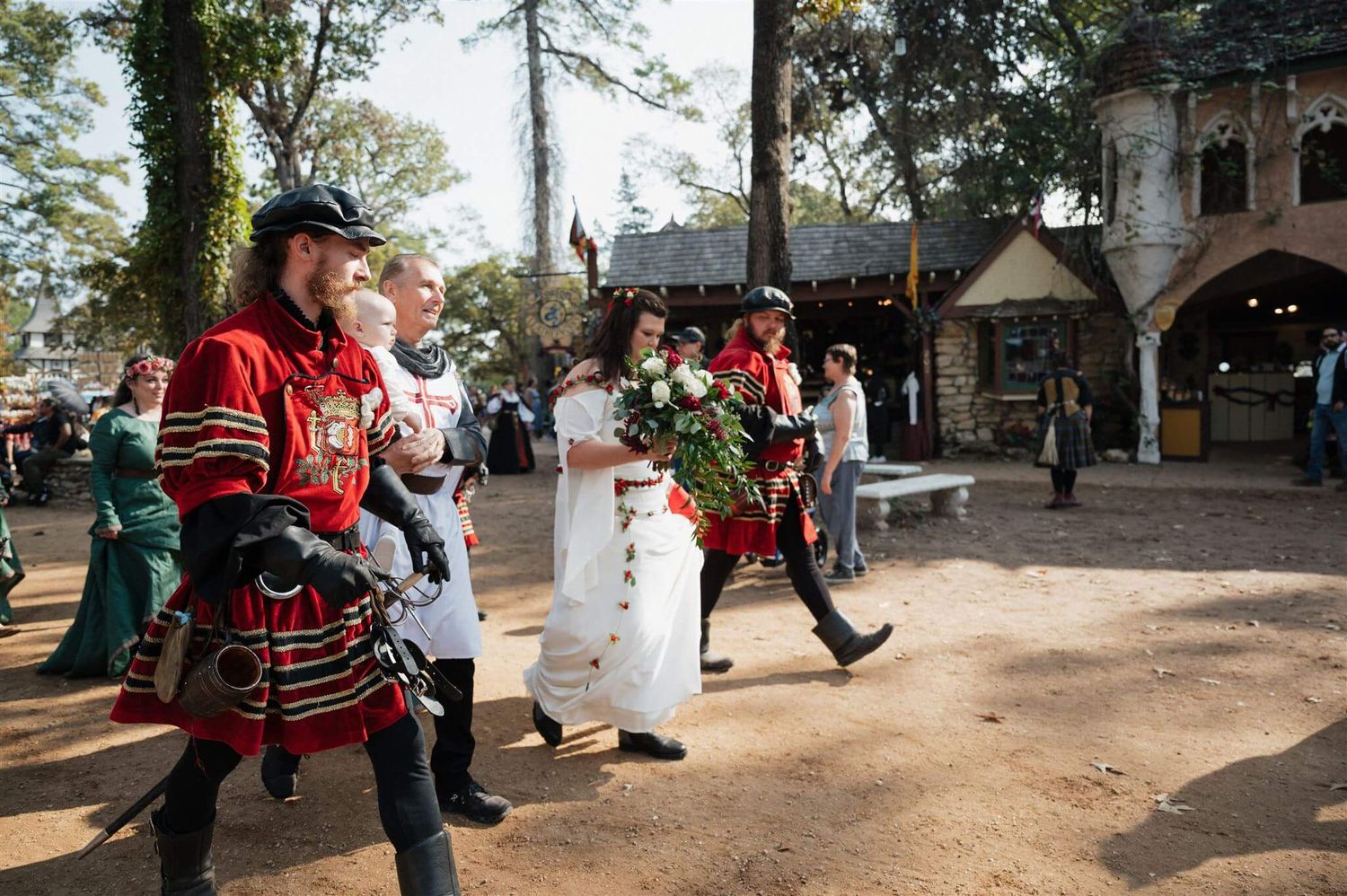 Medieval performers in red and white costumes entertain visitors at a Renaissance faire outdoor marketplace.