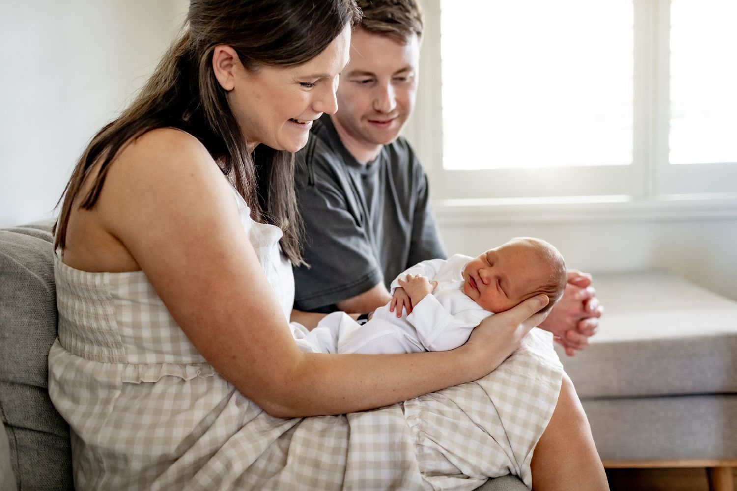 Parents share a joyful moment with their newborn baby while sitting on a grey couch near a window.