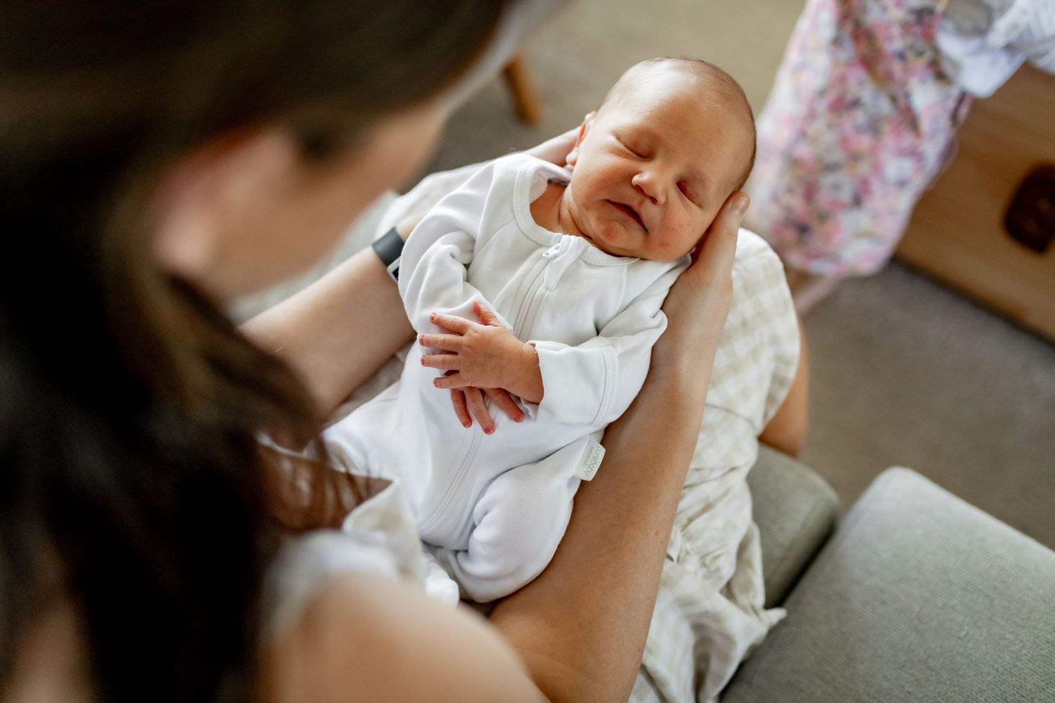 A newborn baby in white clothing smiles while being held on a grey couch.