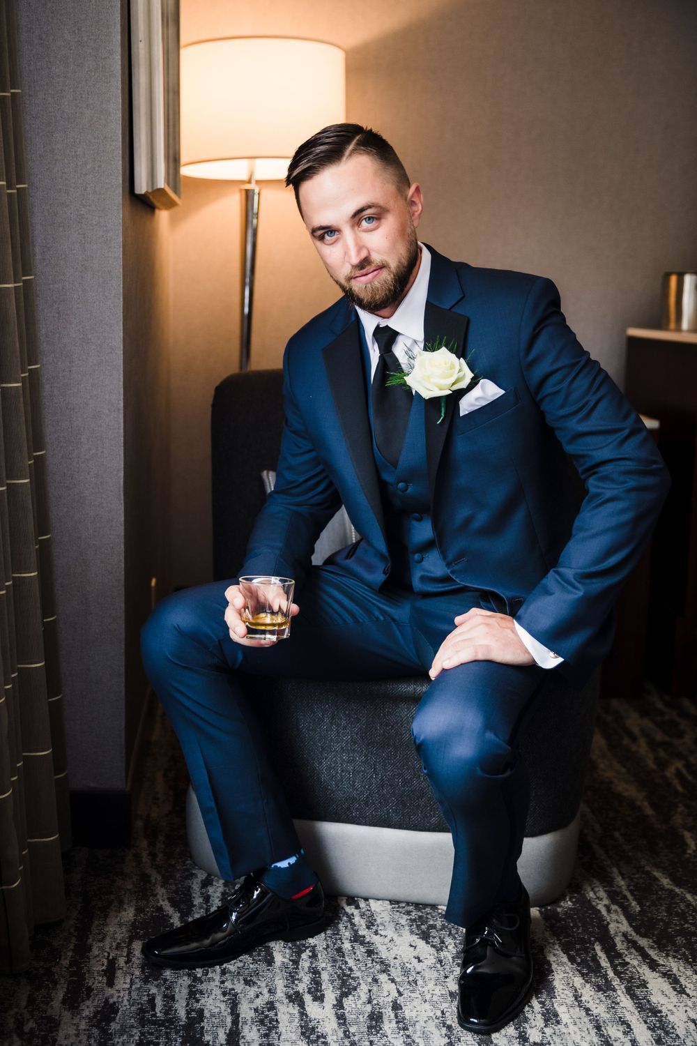 A groom in a navy blue suit sits in a hotel room holding a drink.