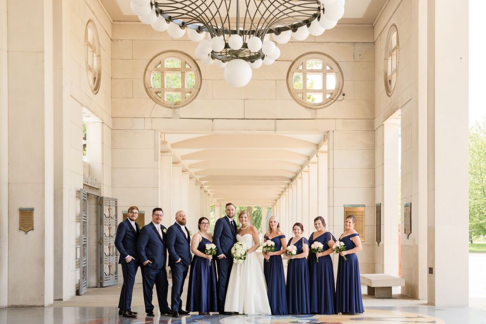 Wedding party in navy and white poses in an elegant columned hallway with chandeliers.