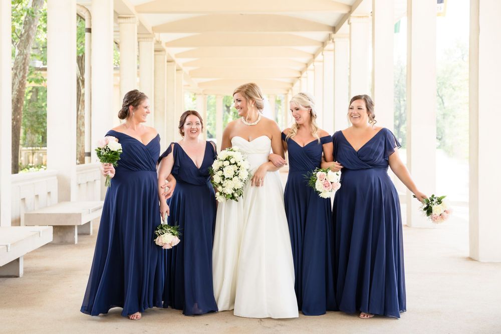 Bridal party in navy blue dresses poses with bride in white gown in columned corridor.