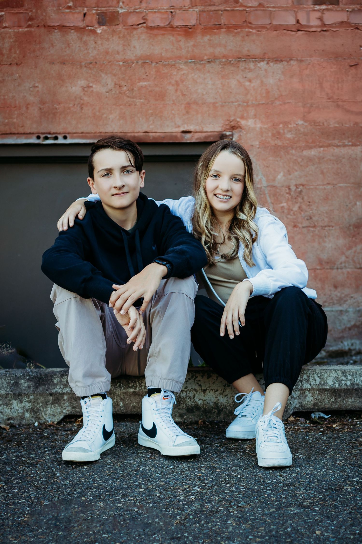 Two people sitting casually against red brick wall wearing sneakers.