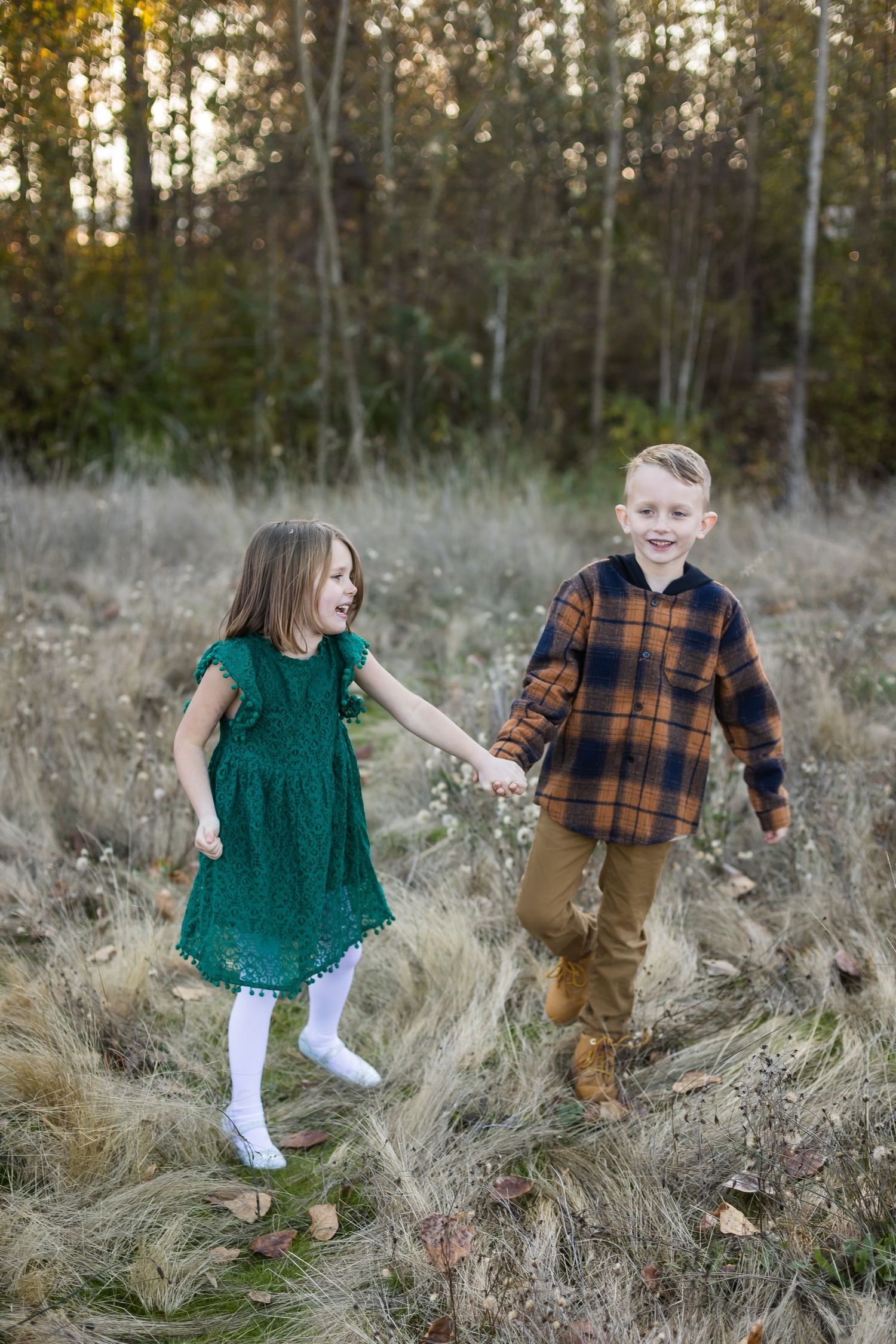 Two children holding hands and playing in a field with brown grass and autumn trees.