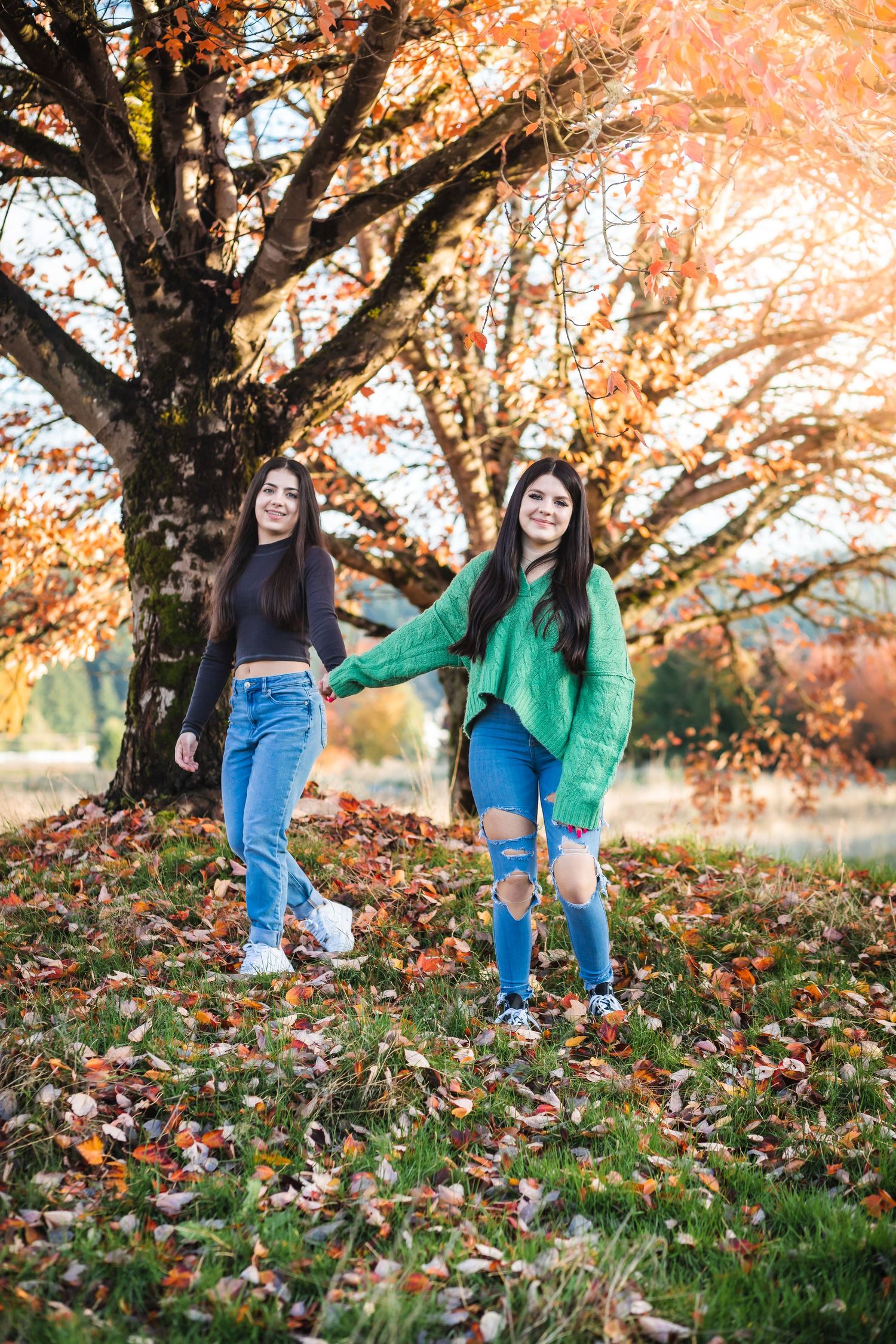 Two friends playing in fallen autumn leaves under colorful tree.