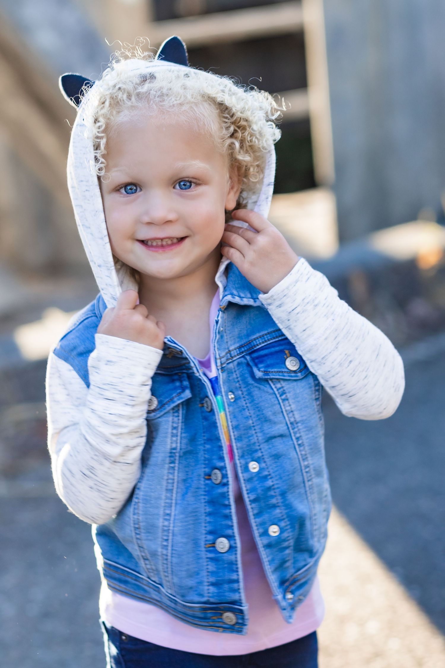 Toddler wearing denim vest with white hoodie sleeves smiles at camera.