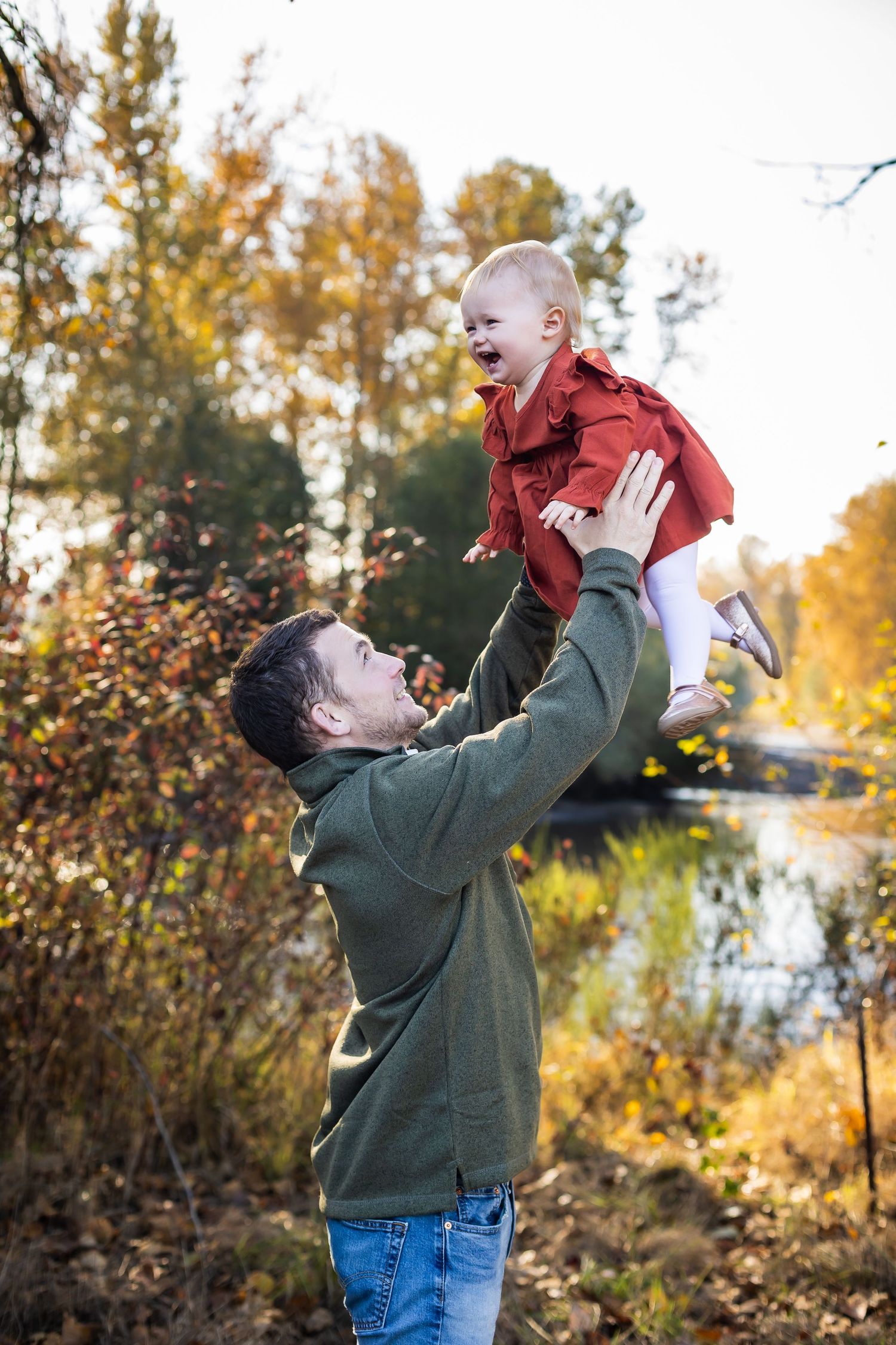 Playful outdoor moment with laughter and lifting in a scenic autumn setting near water.
