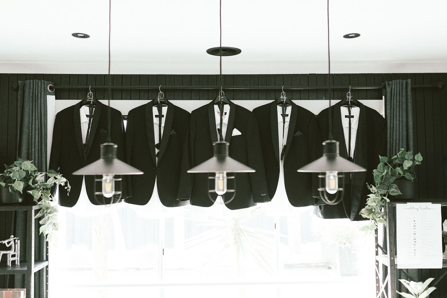 Five black tuxedo jackets hanging in a row with pendant lights overhead.