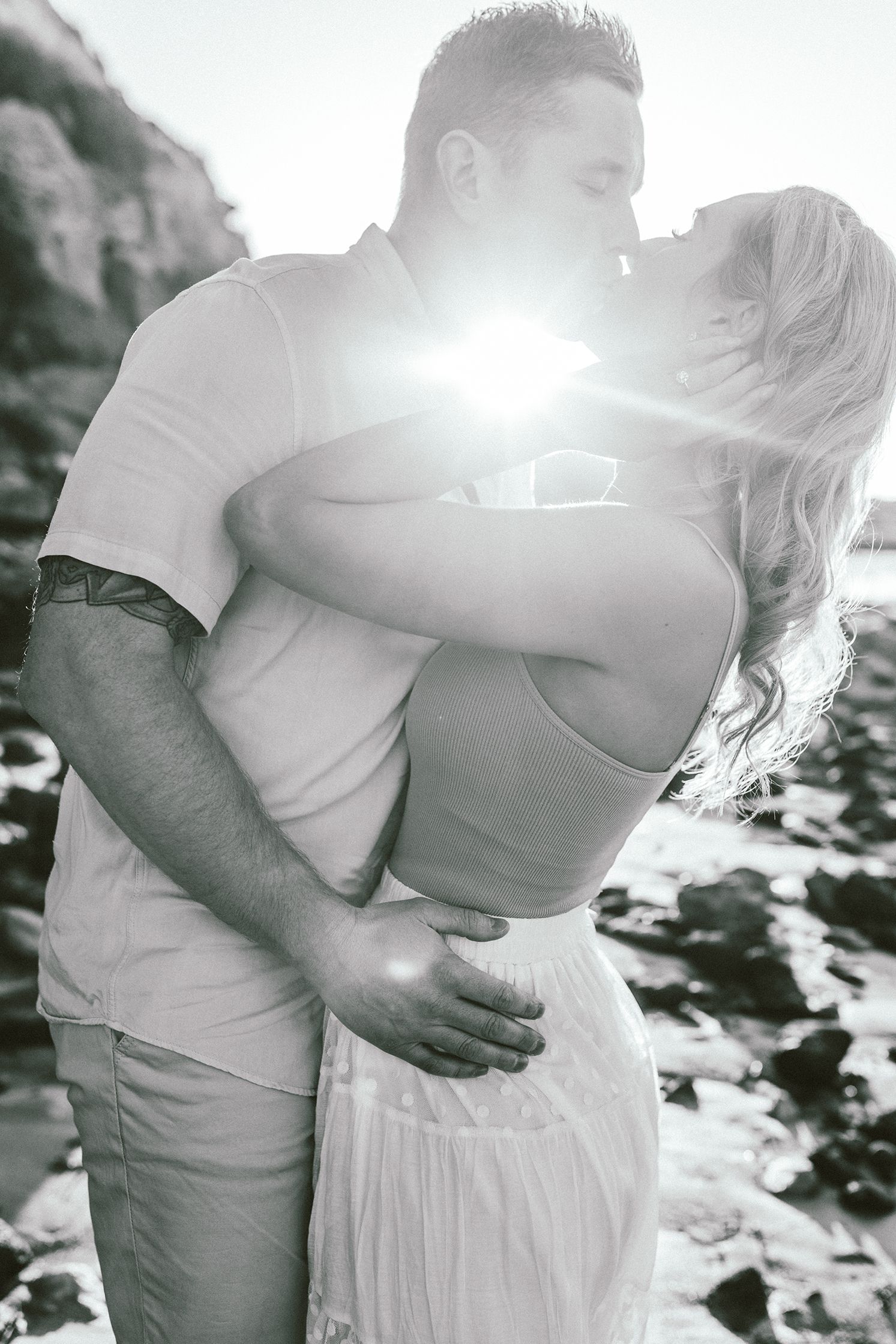 A romantic black and white photograph of a couple embracing on a rocky beach with sun flare.