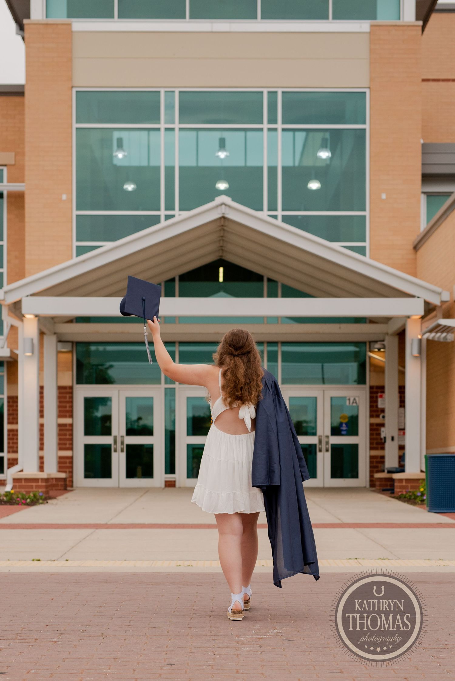 Graduate holds cap high while walking away from modern school building entrance.
