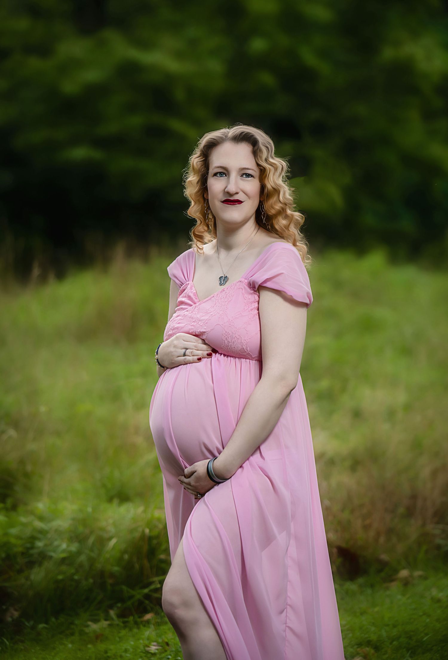 Woman in pink maternity dress cradles baby bump during outdoor photo session.