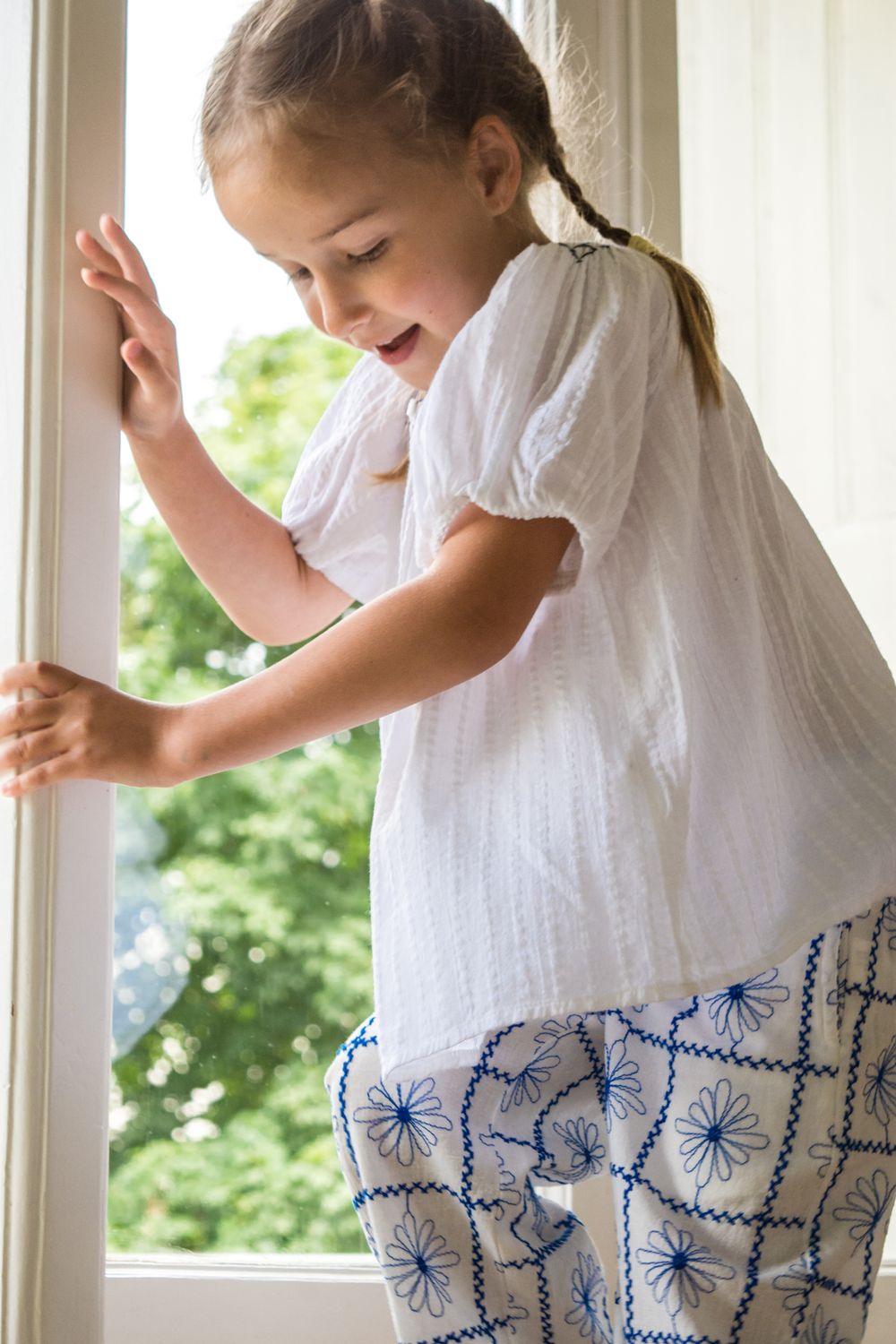 Child in white shirt and patterned pants stands by window with joyful expression on sunny day, Suffolk, UK
