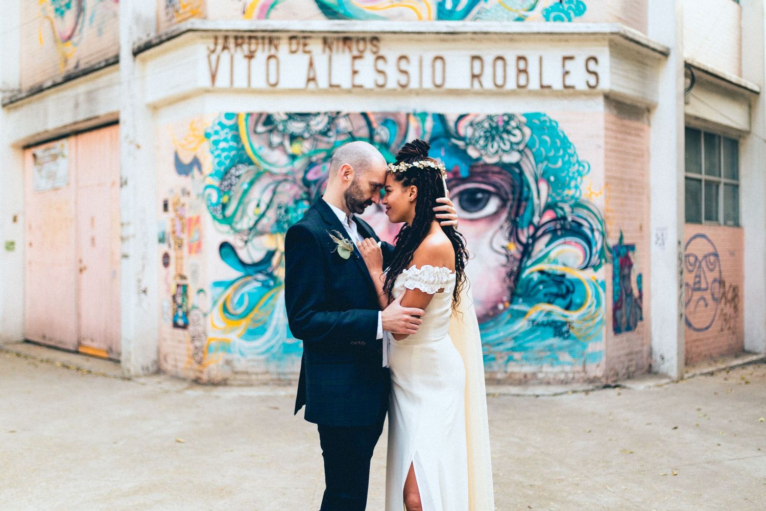 A wedding couple poses in front of vibrant turquoise street art on a corner building.