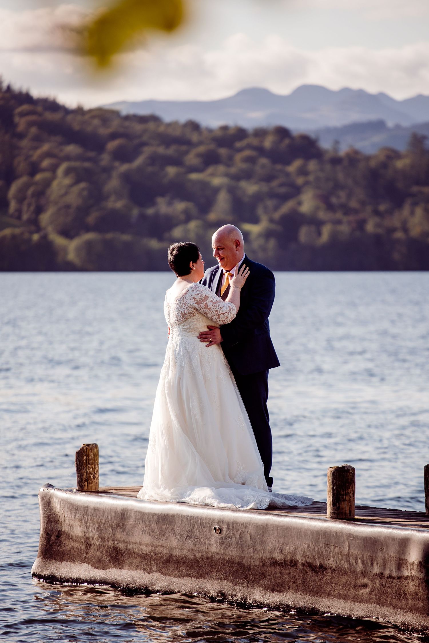 A couple in wedding attire standing on a dock surrounded by mountains and lake waters.