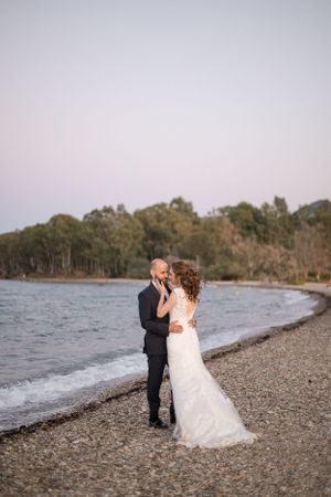 A romantic beach wedding photo of a couple embracing on a rocky shoreline at sunset.