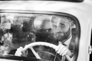 A black and white portrait of someone smiling while driving a vintage car.