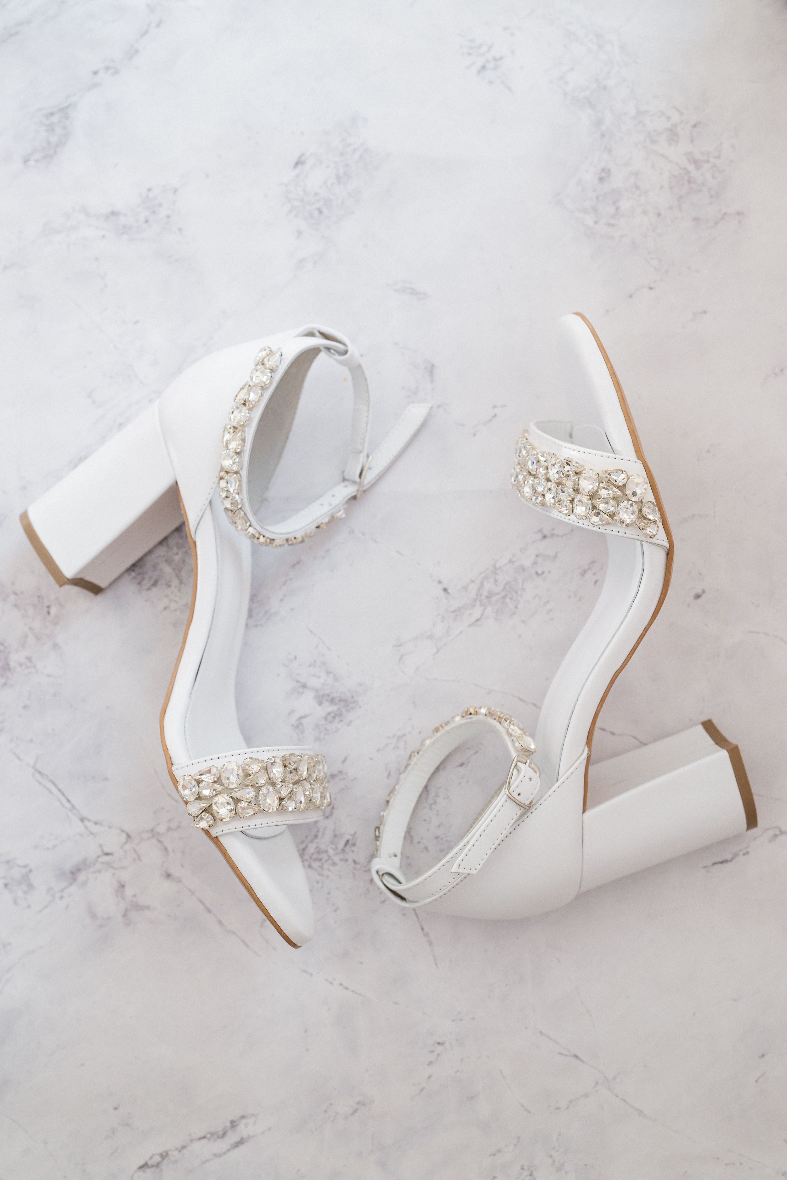 White wedding heels with crystal embellishments and block heel on marble surface.