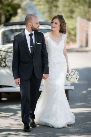 A couple takes a romantic stroll along a sunny path with a classic car in the background.