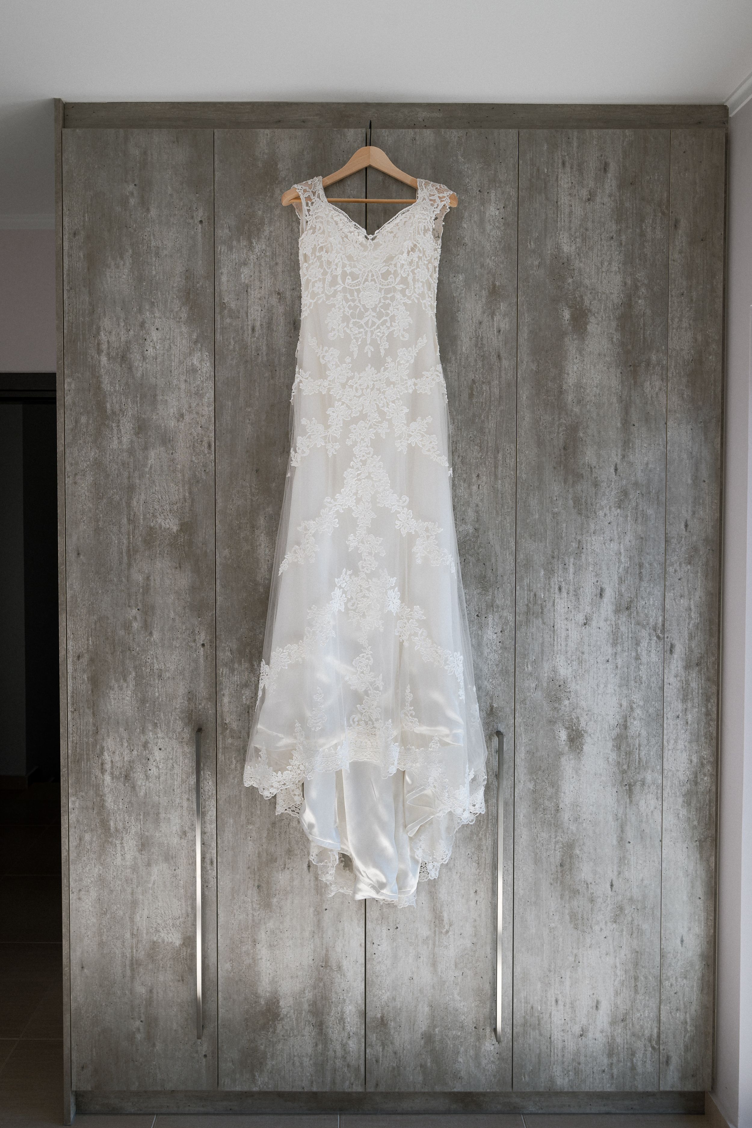 Elegant white lace wedding dress with delicate train hanging on wooden hanger against concrete grey wall.