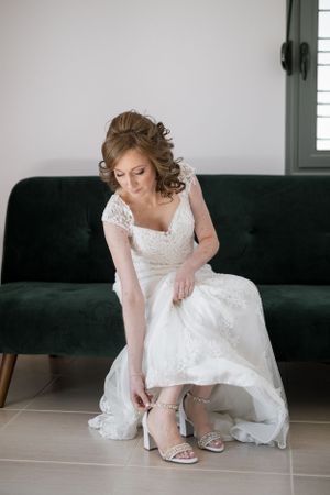 Bride putting on sparkly wedding shoes while seated on emerald velvet couch.