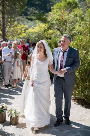 A bride in a white lace dress walks down a sunlit garden path at an outdoor wedding ceremony.