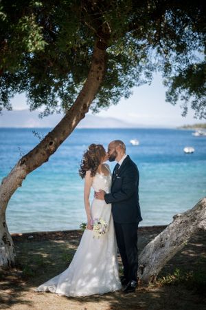 A romantic wedding photo under a tree by the ocean with distant boats visible in the turquoise waters.