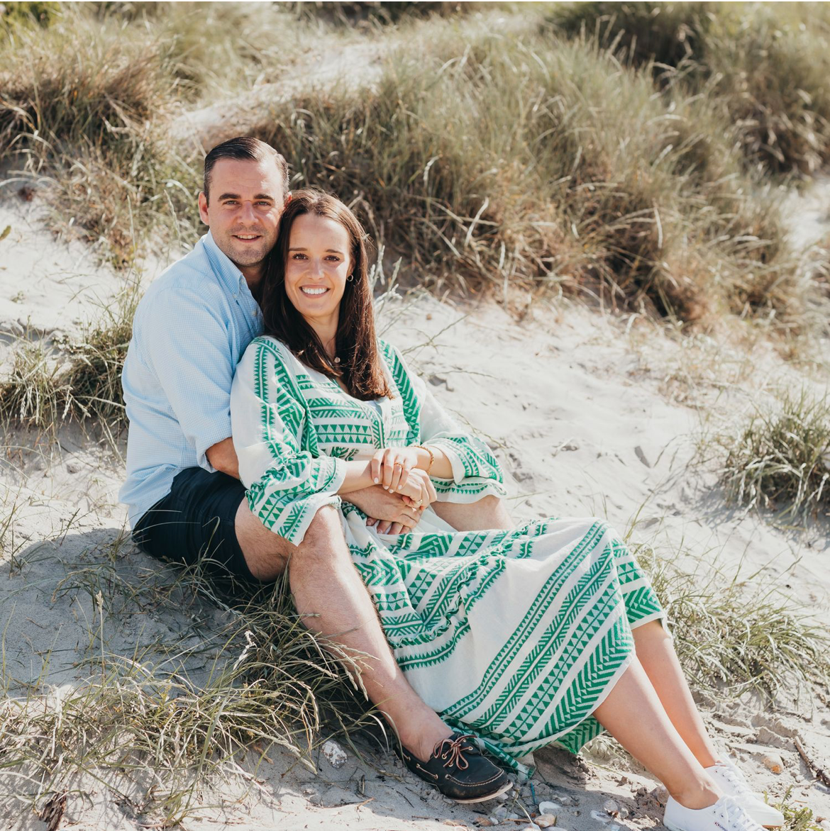 Couple sitting close together on grassy beach dunes with woman wearing patterned green dress during a family beach photoshoot