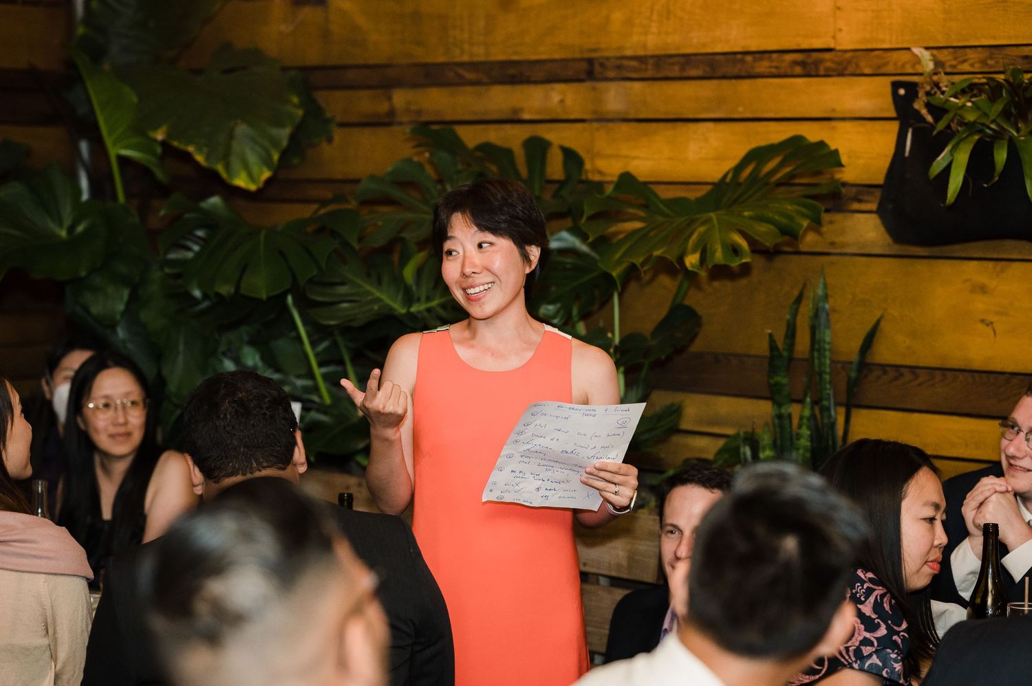 A person in a coral dress gives a speech at a reception dinner venue with wooden walls.
