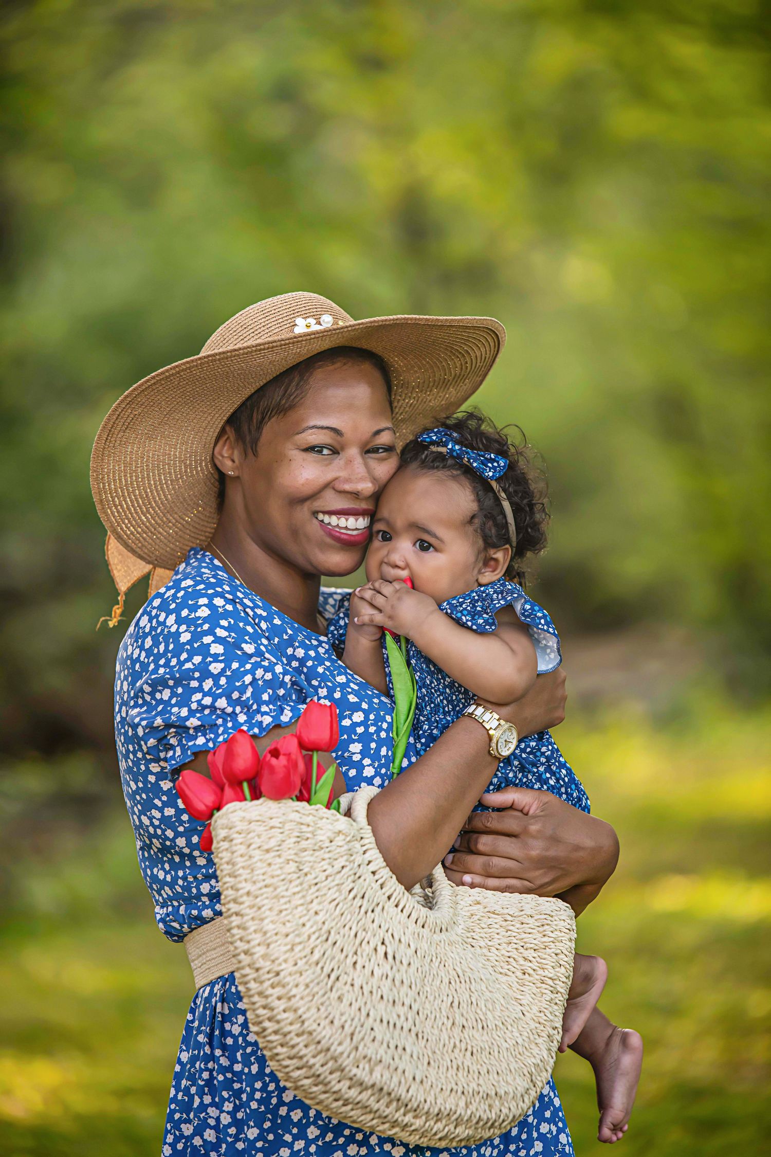 Woman in blue floral dress and sun hat holds smiling baby while carrying straw basket with red tulips in garden setting.