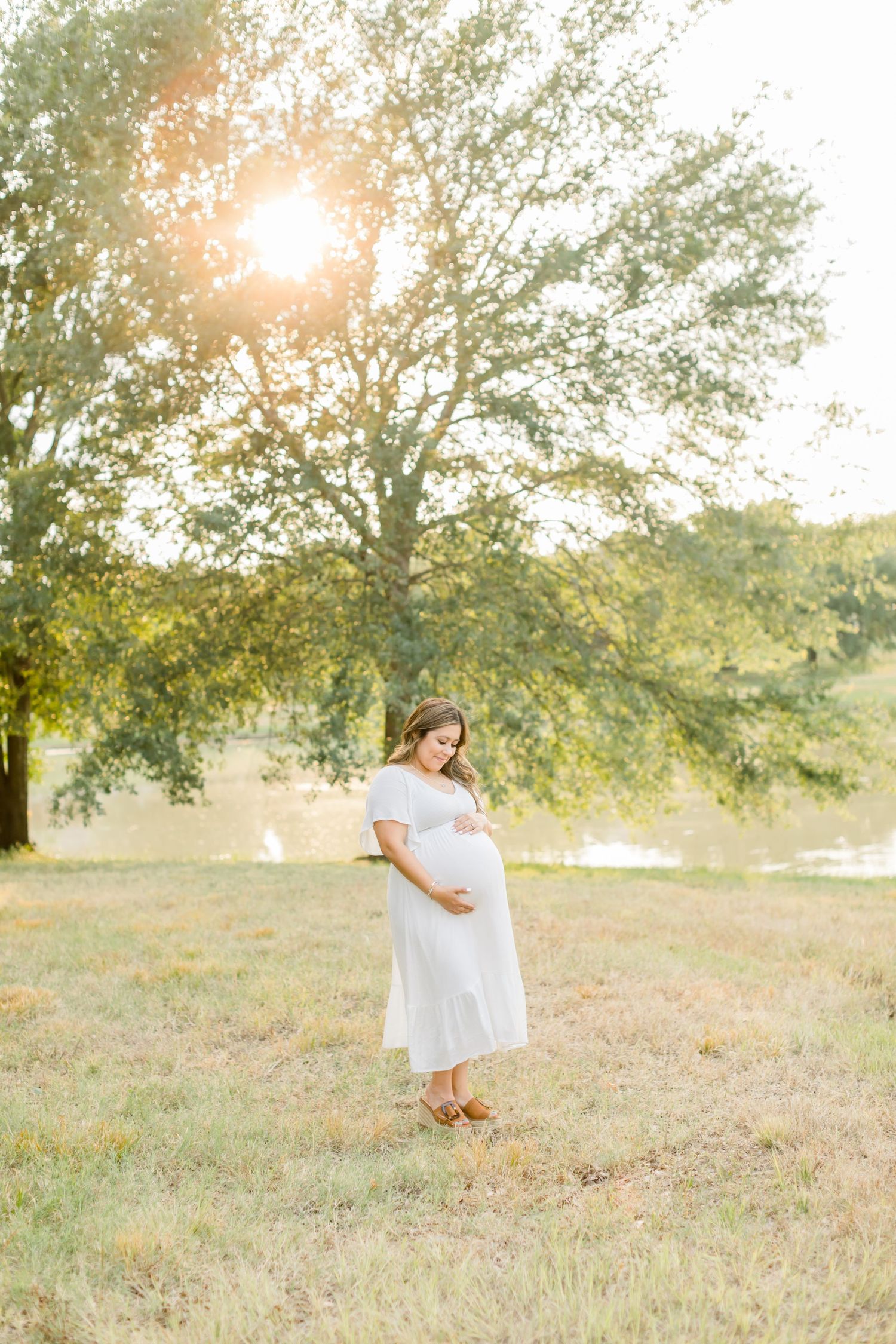 Expectant mother poses in white dress against golden sunset in outdoor field setting.