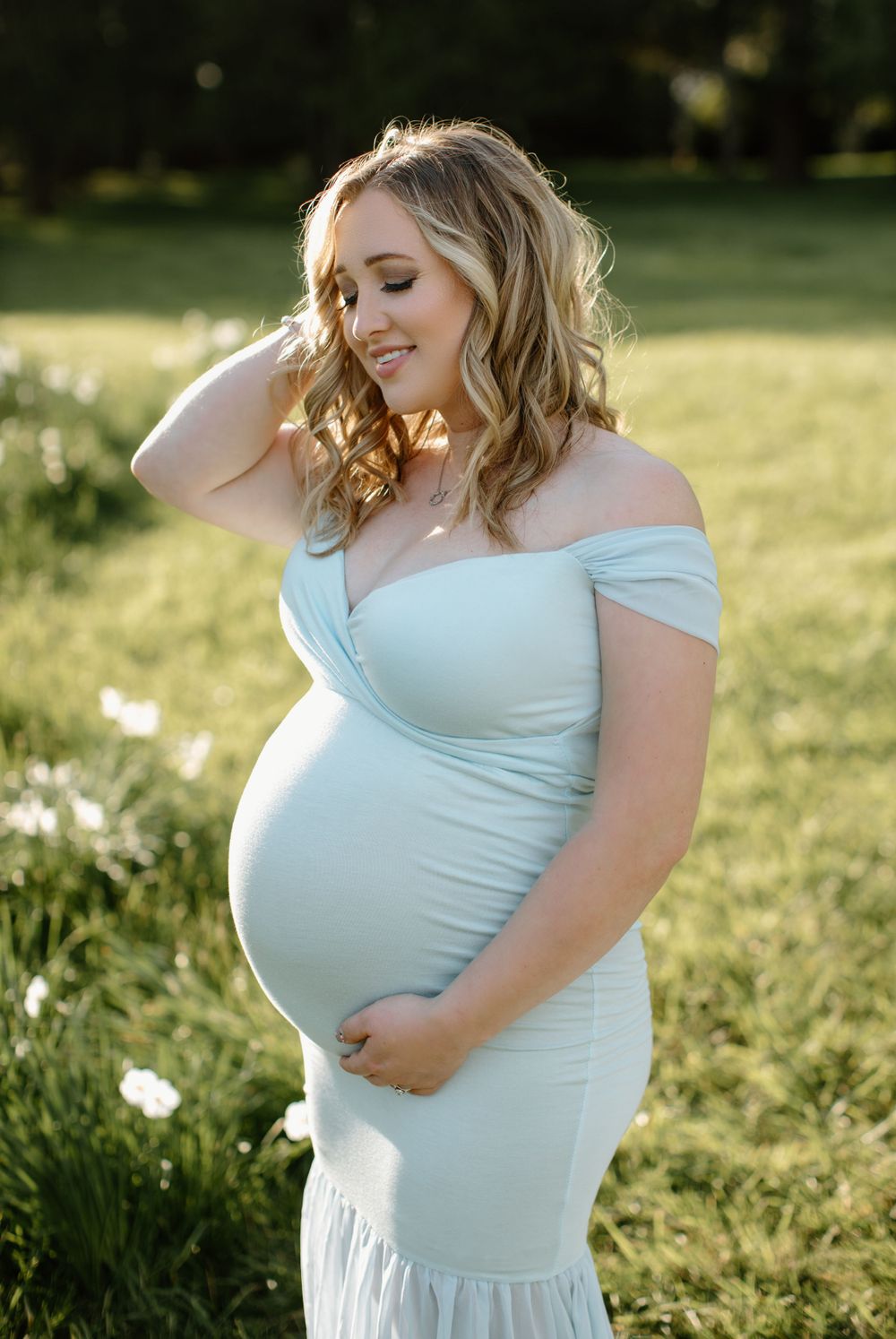 Expectant mother in pale blue dress caresses her baby bump in outdoor setting.