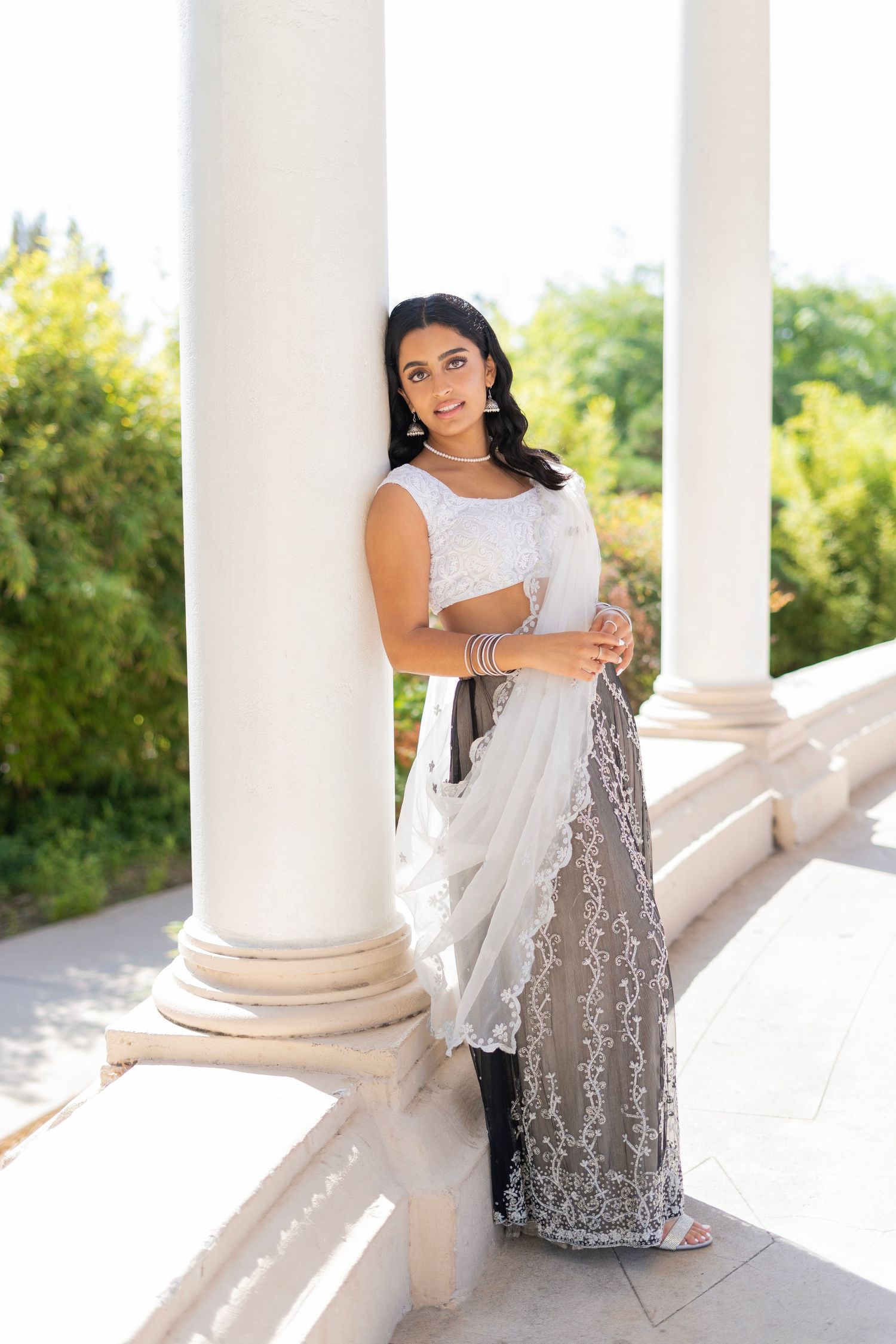 Elegant woman in white and gray traditional saree poses gracefully between classic white columns on a sunlit balcony.