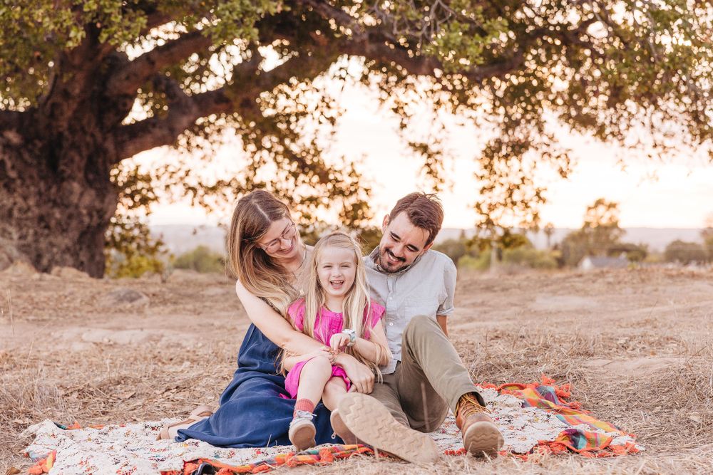 A family sharing a playful moment on a blanket under an oak tree at sunset.
