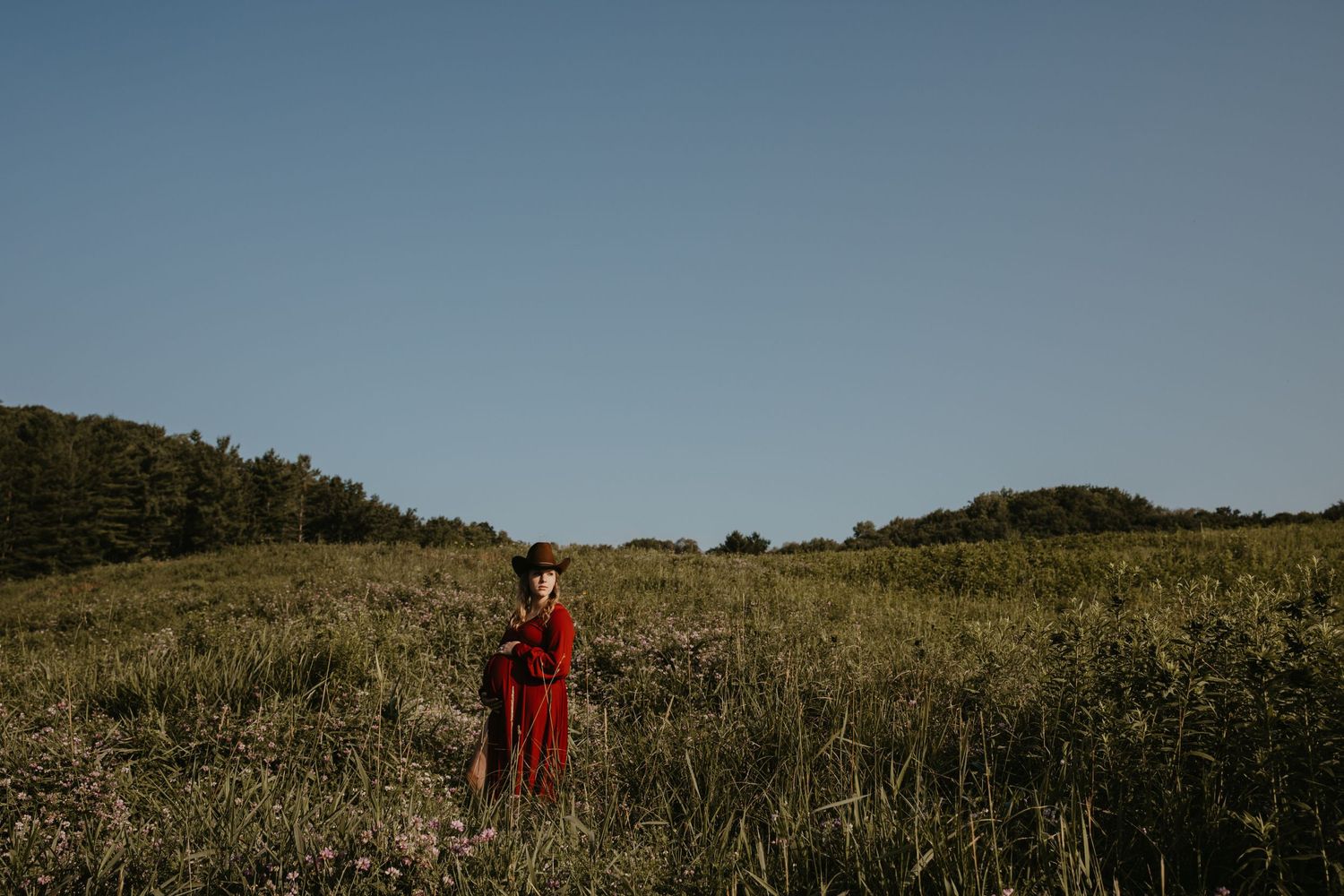 pregnant woman in a long red dress and cowboy hat embraces her belly with her baby, standing in big open grass field with a giant blue sky