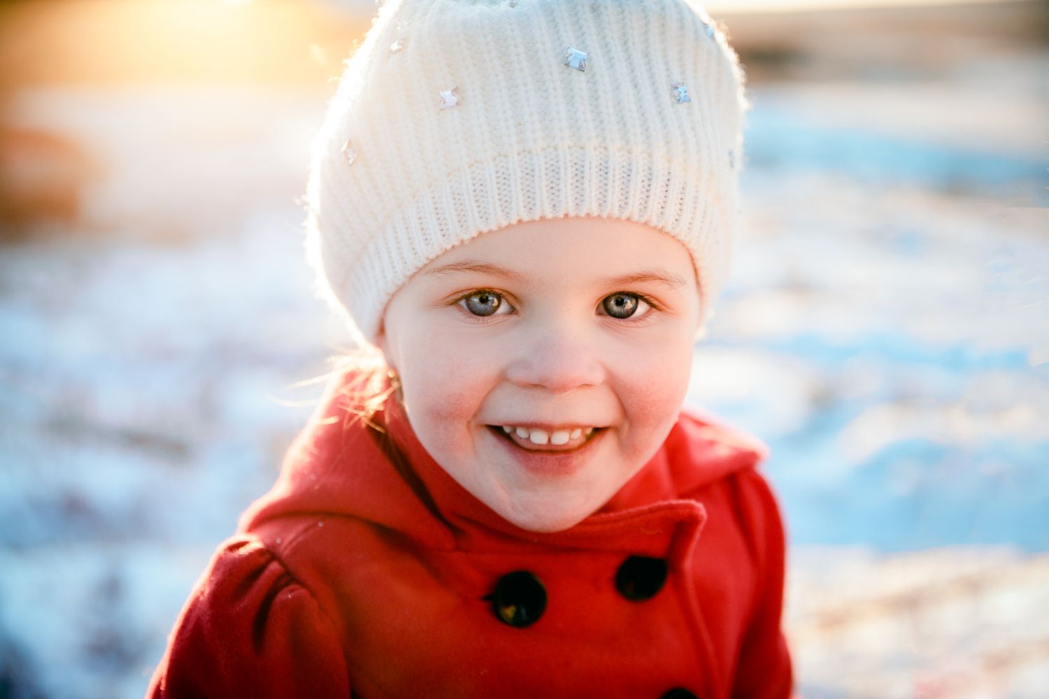 Adorable young girl in a red coat and white knit hat smiles brightly outdoors in a snowy winter setting.