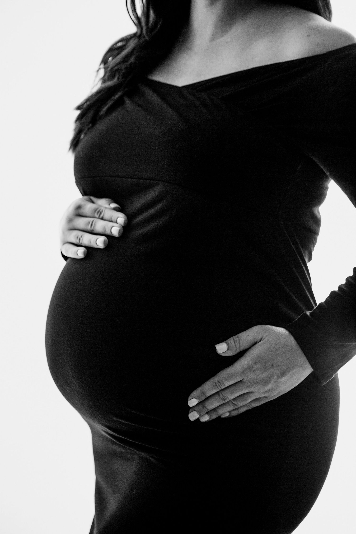 Black and white maternity portrait showing hands cradling pregnant belly in elegant dress.