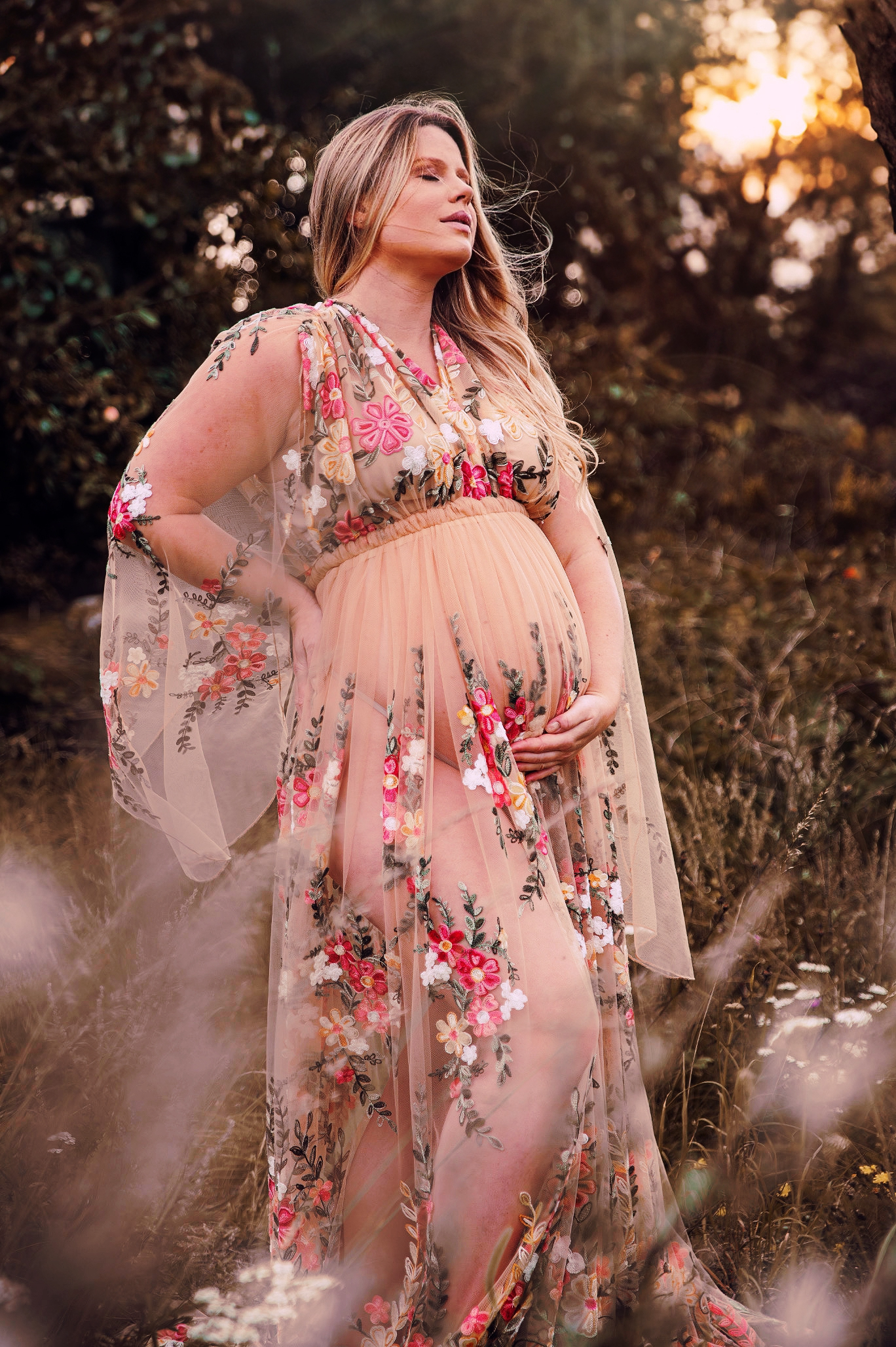 Golden hour maternity photography featuring a romantic sheer dress with pink floral details in a natural setting.