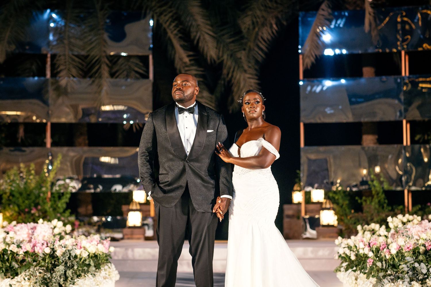 Elegant wedding couple poses for nighttime portrait surrounded by flower arrangements and palm trees.