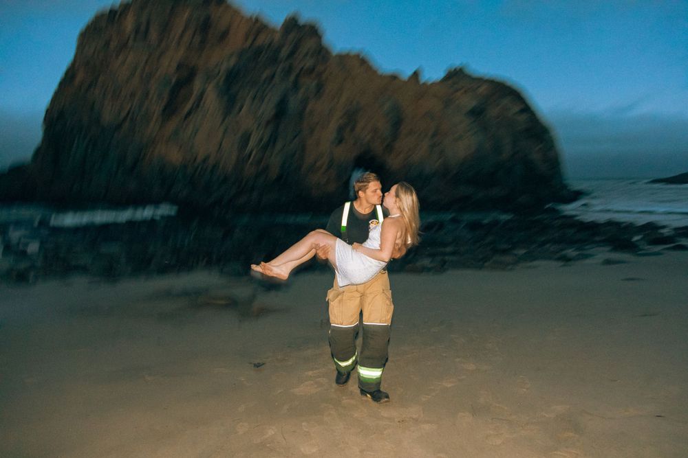 Playful moment captured on sandy beach with towering rock formation at sunset.