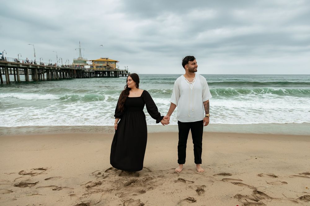 A couple holds hands on sandy beach with pier and ocean waves in the background.