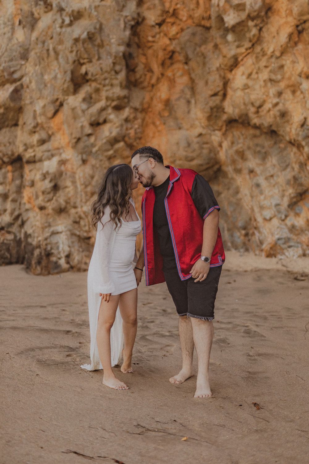 A romantic couple shares an intimate moment on a beach with towering cliffs in the background.