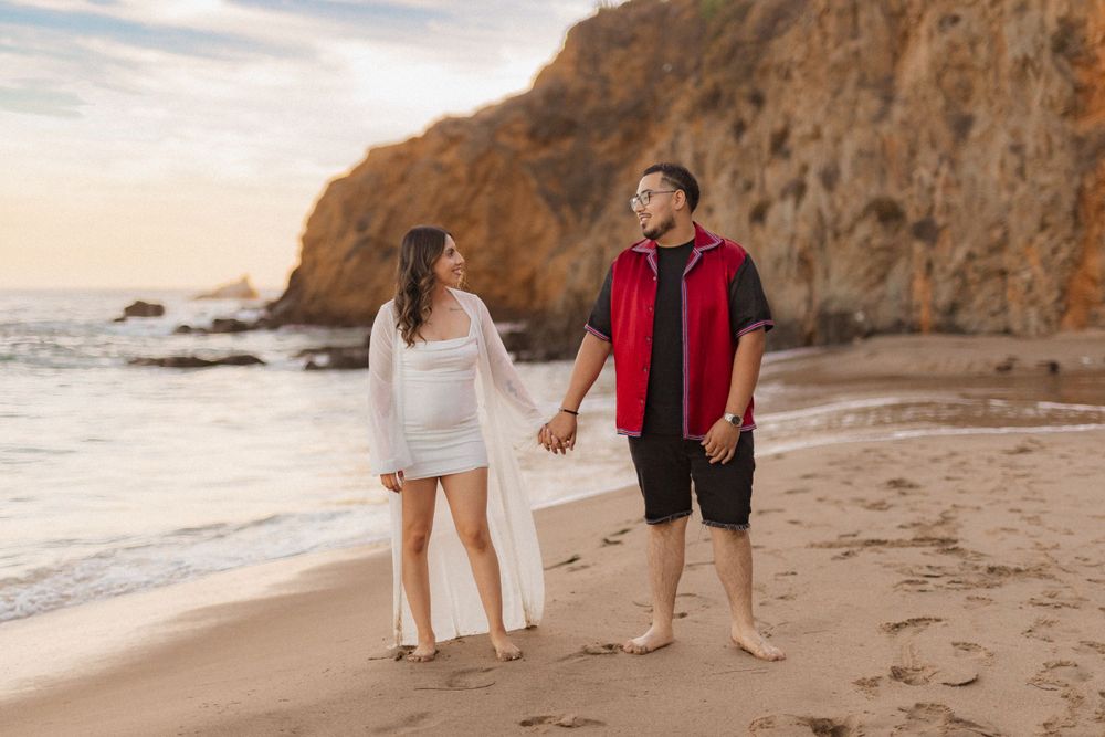 Couple exchanging loving glances while walking on beach with dramatic cliff backdrop.
